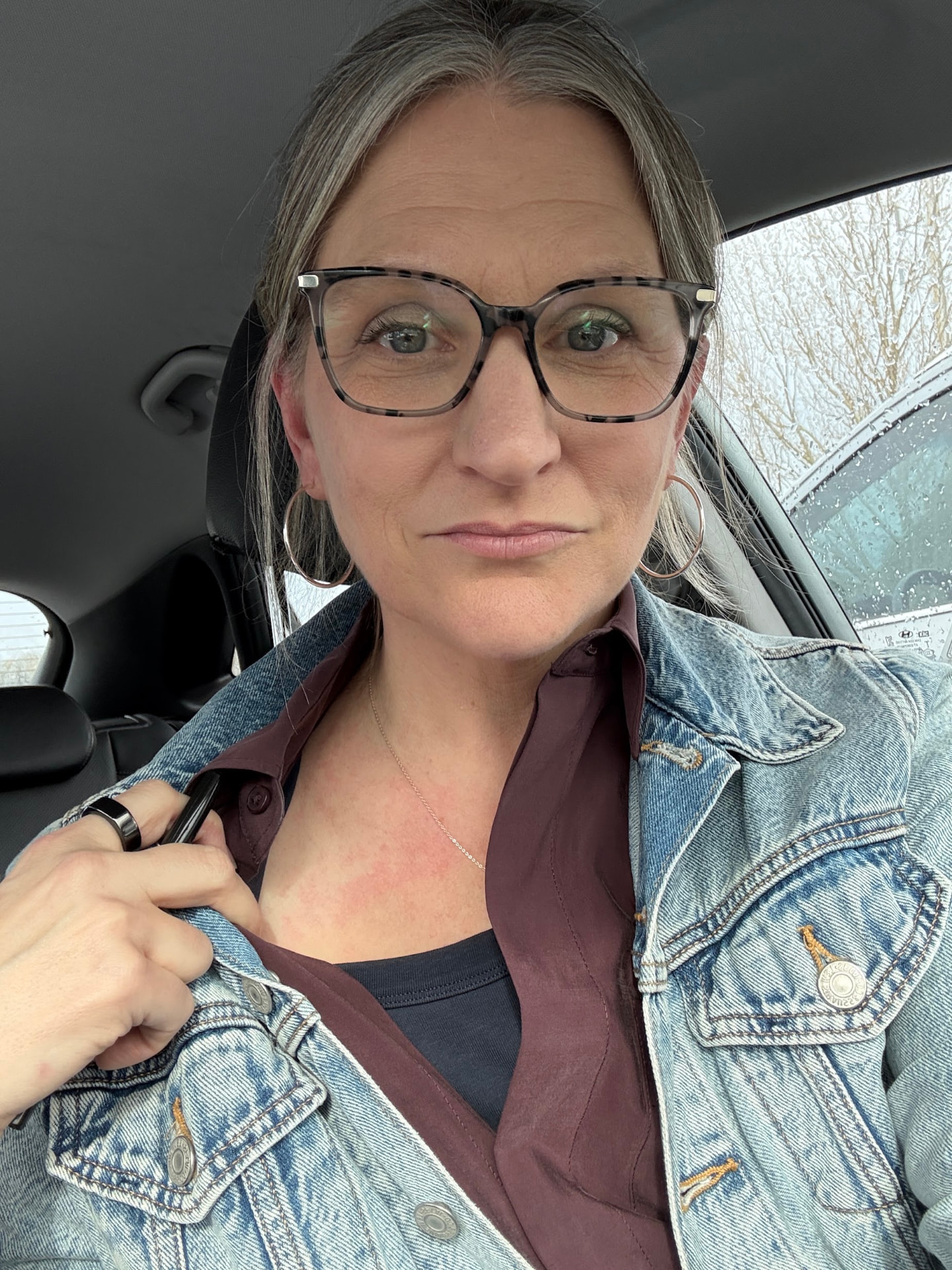 Becky sitting in her car just after the meeting at her daughter's school, wearing glasses, hoop earrings and a denim jacket.